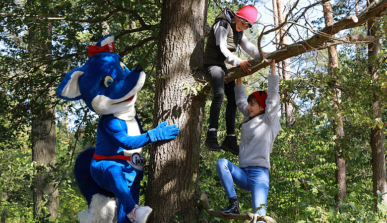 Maskottchen KIKKI der IKK BB steht im Wald neben zwei Kindern, die lachend auf einem Ast klettern