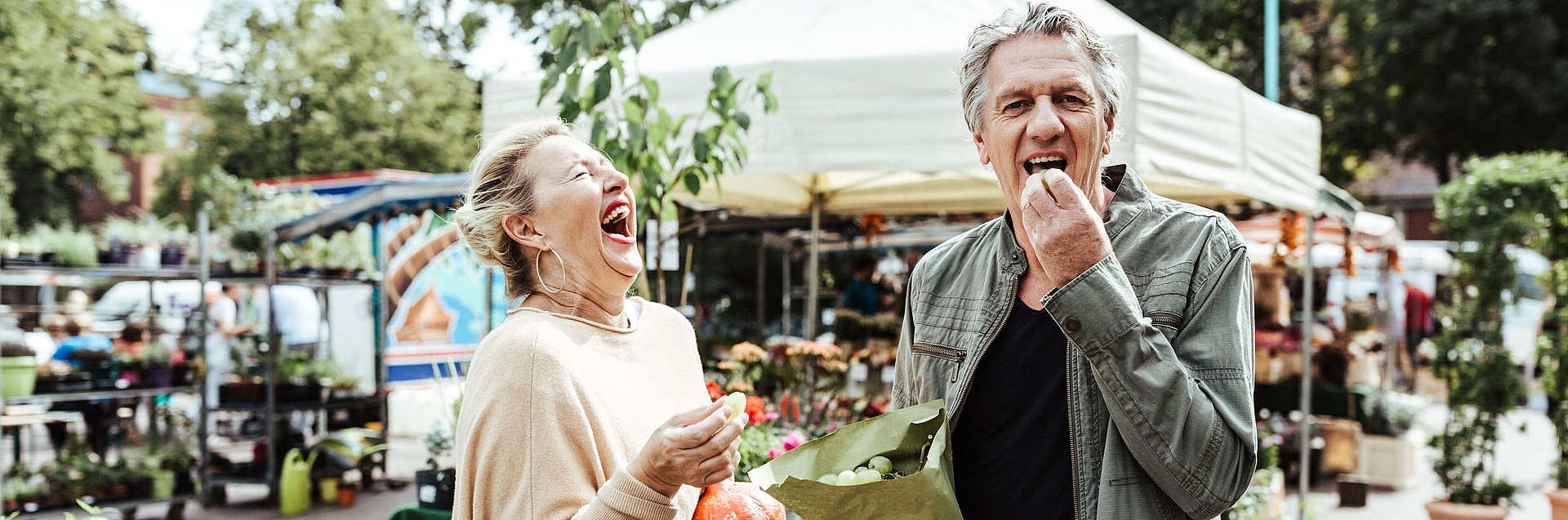 Ehepaar steht lachend auf dem Wochenmarkt und nascht Weintrauben.