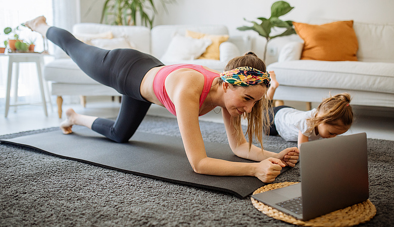 Frau macht eine Yoga-Übung auf einer Matte und schaut dabei auf einen Laptop mit einem Online-Tutorial.