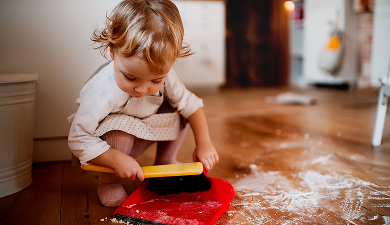 Kleines Mädchen sitzt am Fußboden und fegt mit Handfeger und Schaufel den Fußboden sauber