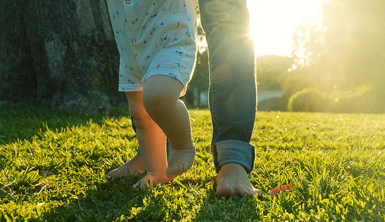 Baby macht erste Schritte auf grünem Rasen, umgeben von Männerfüßen.